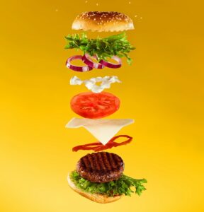 Burger vertically disassembled and each ingredient floating on a mustard coloured background
